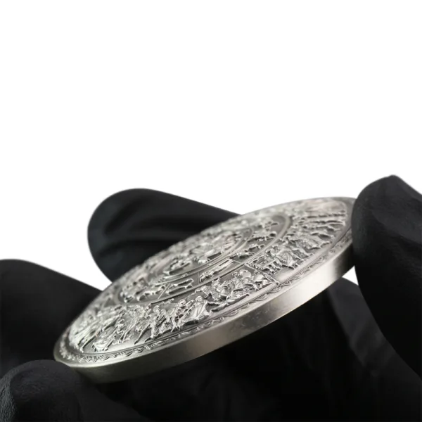 Detailed silver commemorative coin held in black gloves, featuring intricate relief designs on both sides, showcasing historical or cultural motifs.